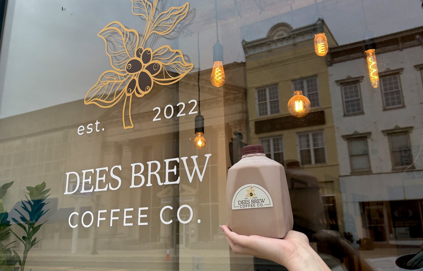 A hand holding a 32 ounce jug of Dees Brew iced coffee up in front of the shop window